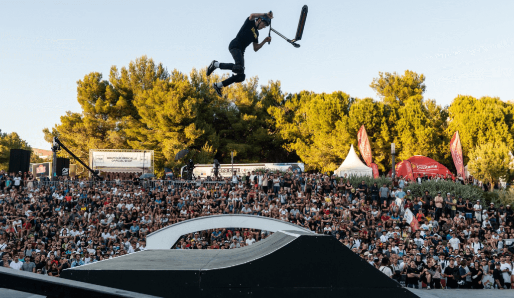 Acrobatie d'un sportif qui fait de la trottinette. Sa pirouette est dans l'air. On voit une foule de personnes derrière lui.